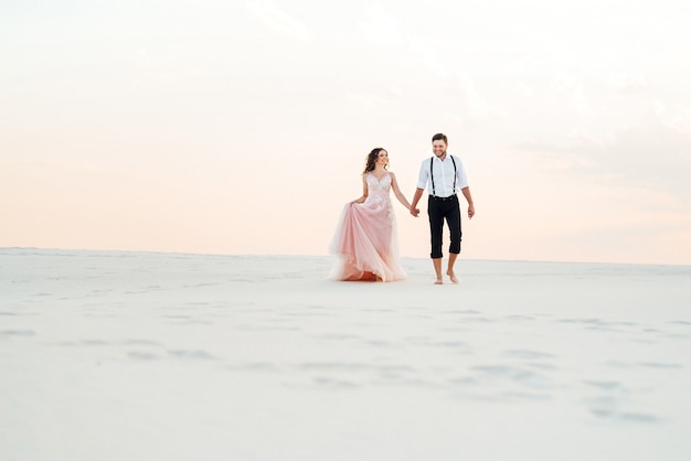 Young couple a guy in black breeches and a girl in a pink dress are walking along the white sand of the desert