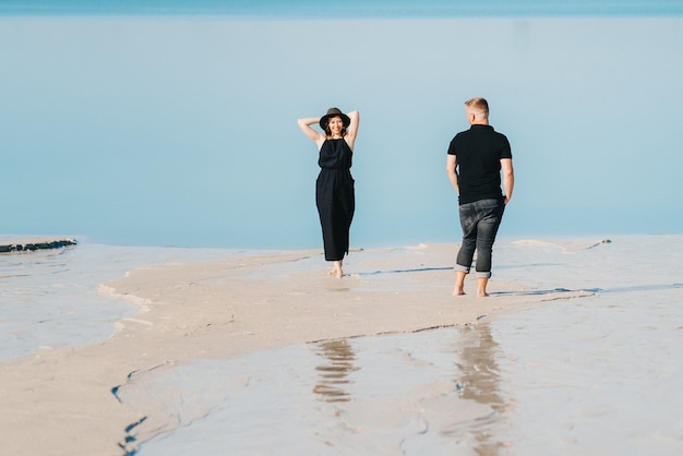 Young couple a guy with a girl in black clothes are walking on the white sand at the edge of blue water