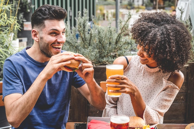 Young couple having fun at restaurant two diverse friends eating burgers at fast food and drinking beer