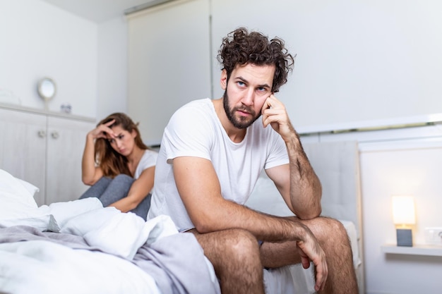 Young couple having a problem. Handsome man is leaning on his hand while sitting sadly on bed, woman is sitting in the background. Head full of thoughts when she is around