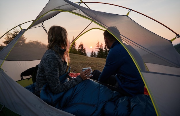 Young couple hikers sitting in camp tent and looking at sunset