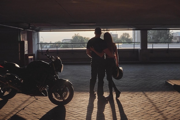 Young couple hugging standing by on a motorcycle in underground parking garage on the sunset