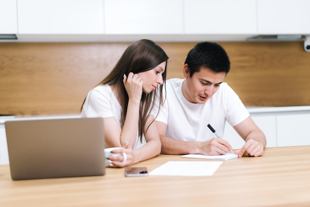 Young couple is talking on kitchen and write in notepad entry note. Confident man explaining to girl information and writes it on paper. Woman smiling listening to partner. Laptop standing on table