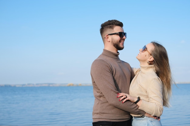 A young couple in jeans sweaters and sunglasses stands on the beach near the lake and hugs