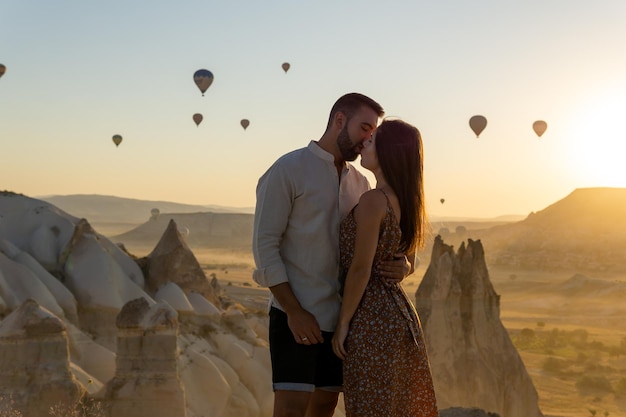 Young couple kissing very much in love in the foreground looking at the typical hot air balloons of the cappadocia area flying at sunrise