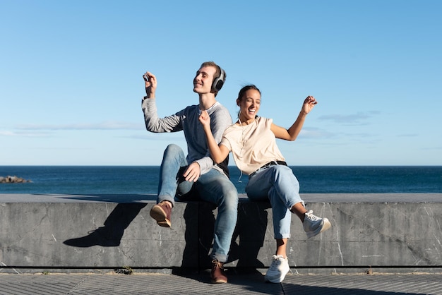 A young couple listening to music and dancing sitting on a stone seat at the seaside in a sunny day