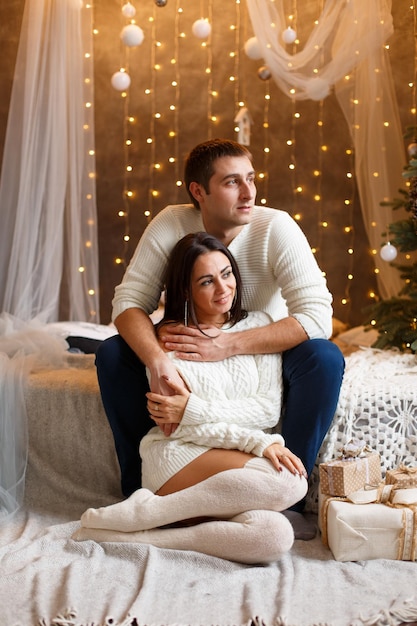 Young couple in love man and woman are sitting by bed hugging kissing in bedroom near christmas tree...