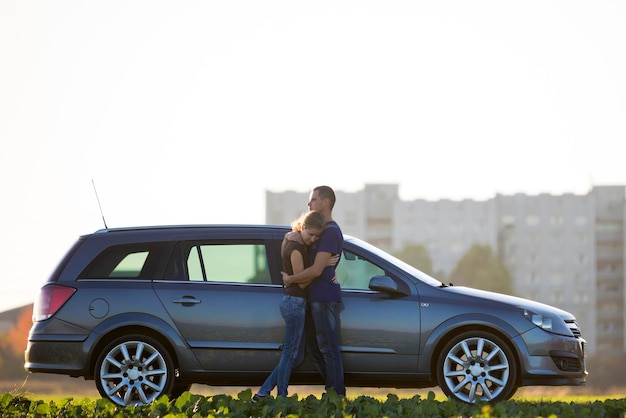 Young couple in love slim attractive woman with long ponytail and handsome man standing together at silver car in green field on blurred apartment building and clear sky copy space background