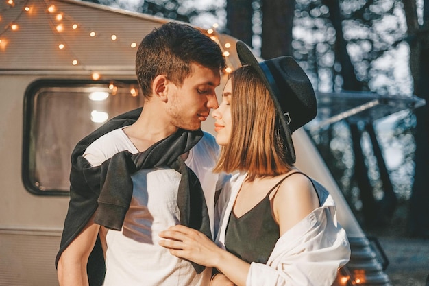 Young couple in love in stylish fashionable outfit romantic weekend wedding trip against the background of camper in woods forest road trip