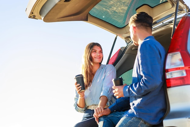 Young couple man and woman traveling together by new car having stop for drinking coffee in a wheat field at sunset