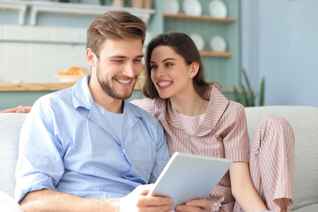 Young couple in pajamas watching media content online in a tablet sitting on a sofa in the living room.