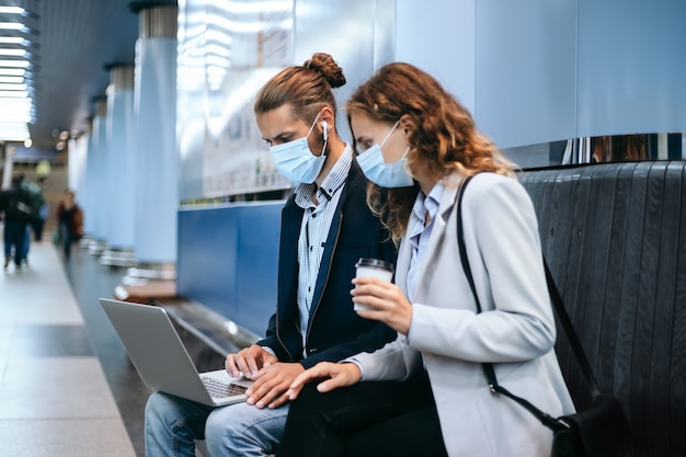 young couple in protective masks using a laptop on the subway platform