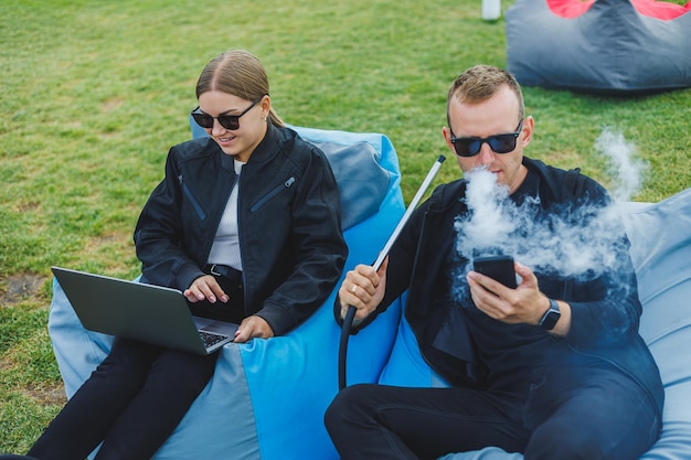 A young couple relaxes in the park and smokes hookah A man and a woman sit on chairs in nature and smoke a hookah Family vacation