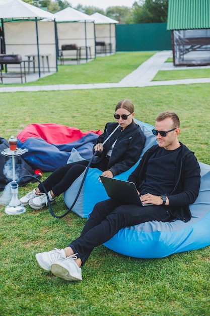 A young couple relaxes in the park and smokes hookah A man and a woman sit on chairs in nature and smoke a hookah Family vacation