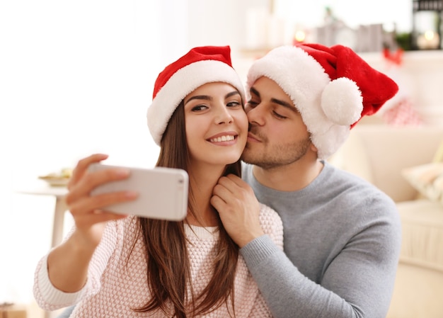 Young couple in Santa Claus hats making selfie indoors