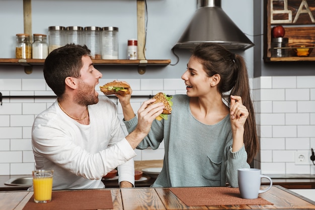 Young couple sitting at the kitchen during breakfast at home, feeding each other with sandwiches