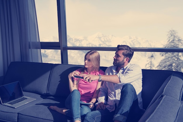 Young couple sitting on sofa near the window watching television together at luxury home