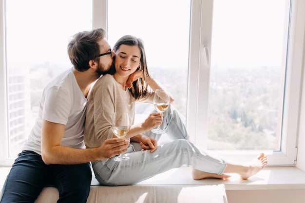 Young couple sitting on windowsill hugging and drinking wine