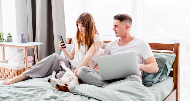 Young couple spend morning in the bed with gadgets and cute dog. Beautiful woman and man looking at laptop and smartphone. Sunny family morning with pet and technologies