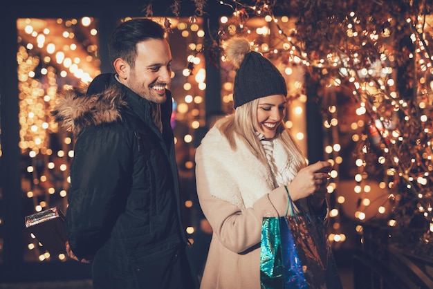 Photo young couple standing in shopping mall with bright shop window in background. a beautiful woman having fun while looking at smartphone.