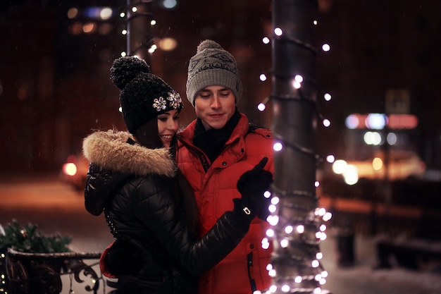 Young couple in sweaters and caps embrace on the winter street. In love in a garland.