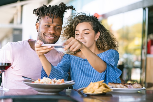 Young couple using a mobile phone and taking photos to the food while having lunch together at a restaurant.