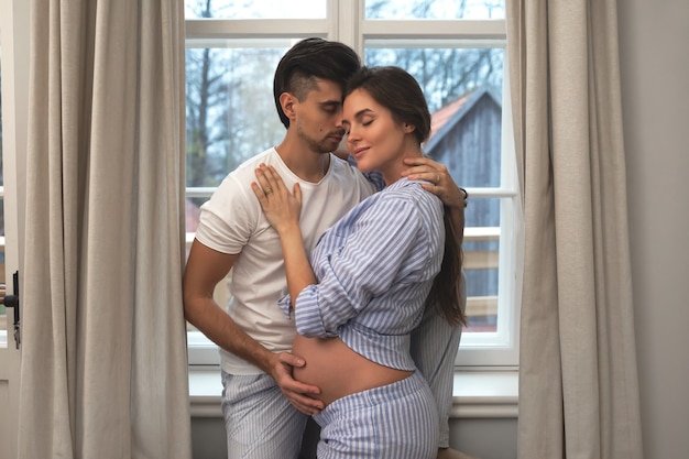 Young couple waiting for a baby. Husband and his pregnant wife in embrace beside the window.