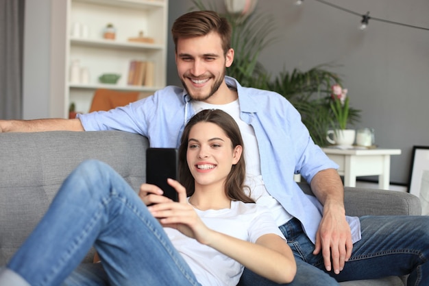 Young couple watching online content in a smart phone sitting on a sofa at home in the living room.