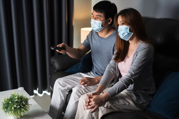 Young couple watching TV on sofa and wearing medical mask to protect coronavirus (Covid-19)