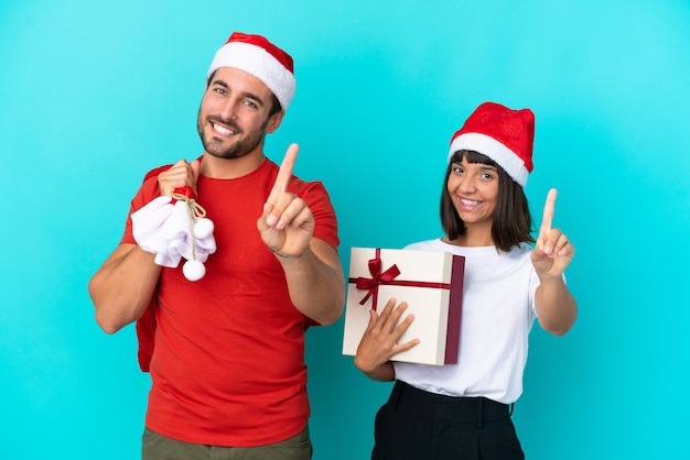 Young couple with christmas hat handing out gifts isolated on blue background showing and lifting a finger