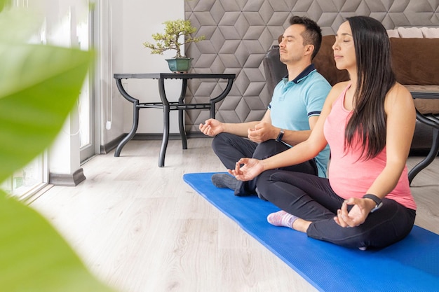 Young couple with pregnant woman meditating in a modern living room at home in front of the window