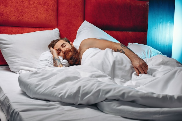 Young   and courageous guy with a long mustache and beard is lying on a white bed and sleeping.