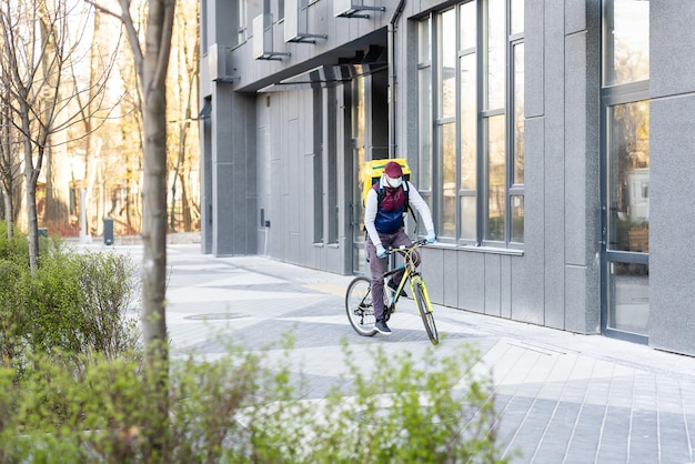 Young courier in medical mask delivering food with yellow thermal backpack, riding a bicycle in the city. Food delivery service concept.