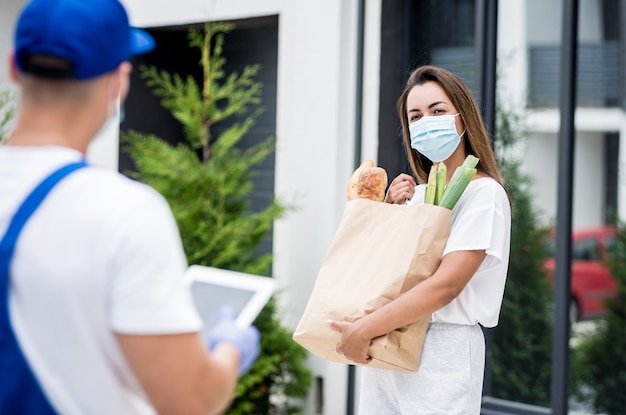 Young courier wearing a protective mask and gloves delivers goods to a young woman during quarantine