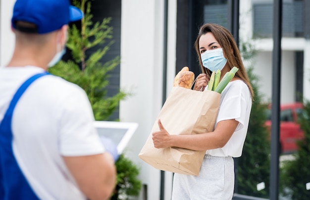 Young courier wearing a protective mask and gloves delivers goods to a young woman during quarantine