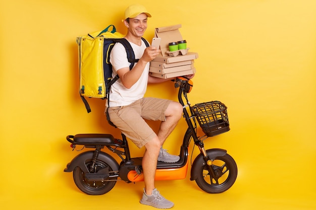 Young courier in white t shirt pizza delivery man with thermo backpack and phone on moped isolated on yellow background Fast transport express home food delivery Online order