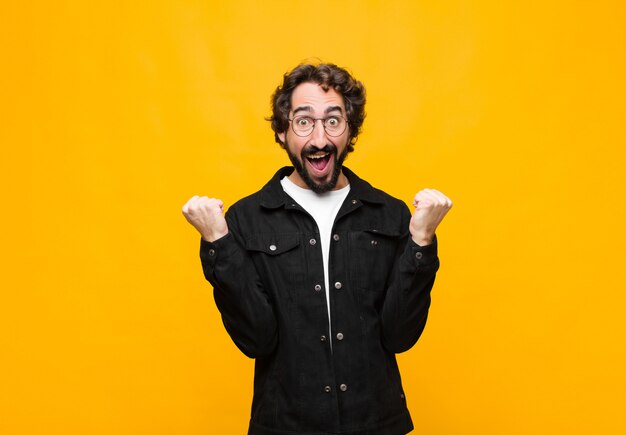 Young crazy handsome man feeling shocked, excited and happy, laughing and celebrating success, saying wow! against orange wall