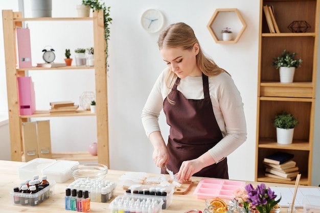 Young creative blonde woman in brown apron chopping soap mass on wooden board while working in studio