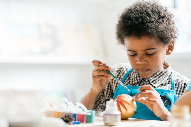 Young creative boy with paintbrush painting golden Christmas toy ball at lesson while preparing for holiday