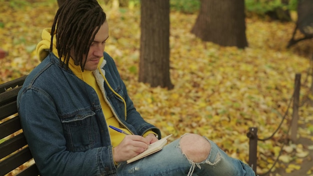 Young creative man with dreadlocks drawing writing in notebook sitting on park bench in golden autumn