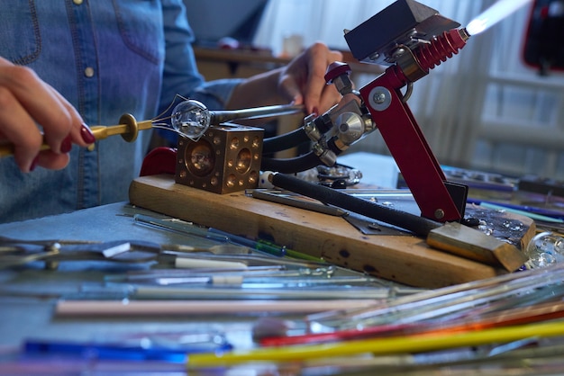 Young creative woman sitting by table in workshop in front of working burner and fixing small glass workpiece with help of handtool