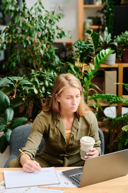 Young creative woman with long blond hair having coffee and making notes