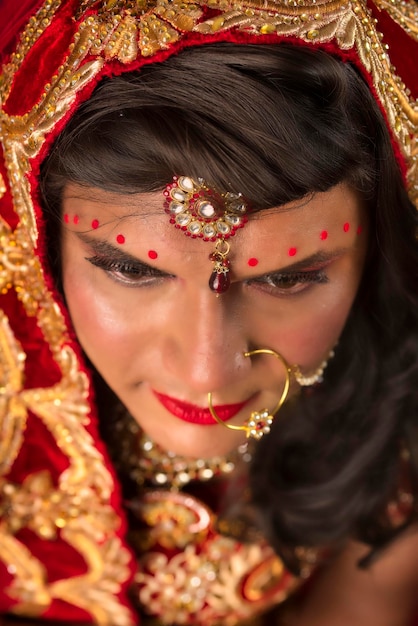 Young crossdresser man wearing Indian wedding clothes of a bride with bridal makeup and posing on grey background looking fashion and glamorous