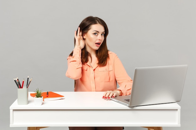 Young curious woman in pastel clothes try to hear you with hand near ear sit work at desk with pc laptop isolated on gray background. Achievement business career lifestyle concept. Mock up copy space.