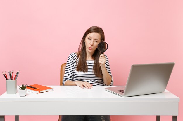 Young Curious woman scrutinizing looking through magnifying glass on pc laptop while sit work on project at office isolated on pastel pink background. Achievement business career concept. Copy space.