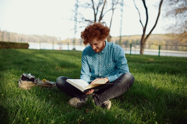 A young curly ginger man riding a book while resting on the grass in the park