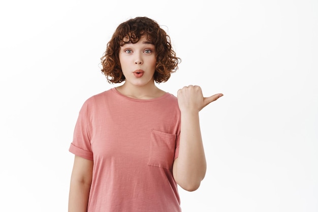 Young curly girl pointing finger right and looking surprised at camera, white background.