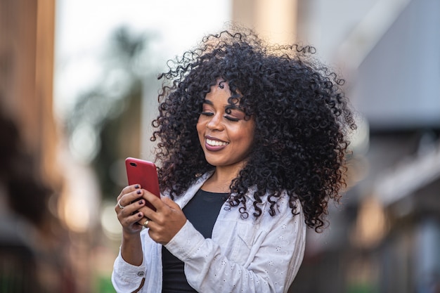 Young curly hair black woman walking using cell phone. Texting on street. Big city.