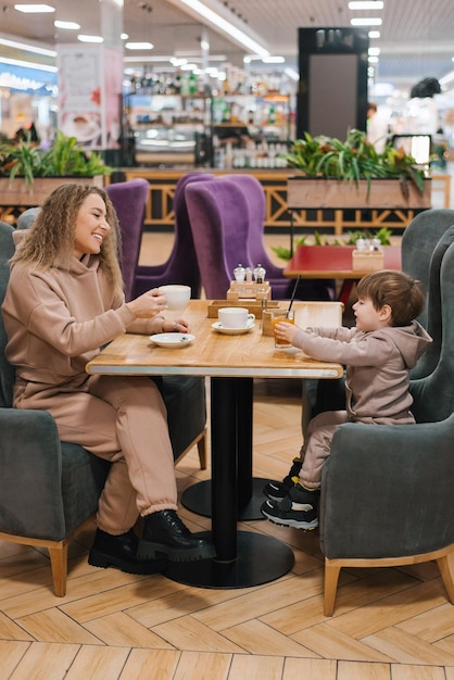 Young curly haired mother and her little son have breakfast or lunch in a cafe sitting at a table