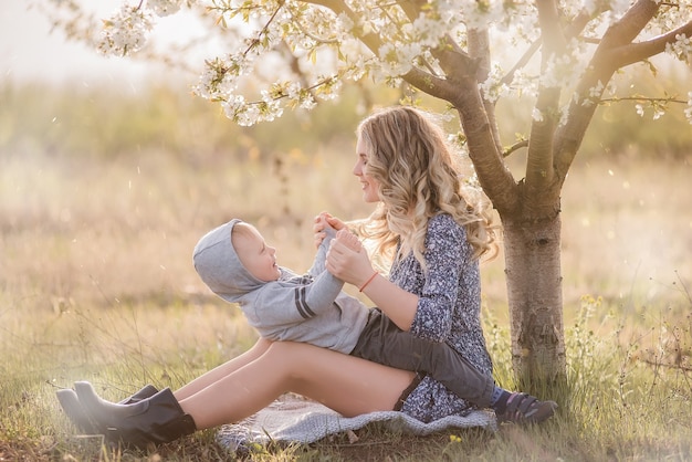 Young curly-haired mother hugs son, sitting in the green grass near blooming white cherry tree